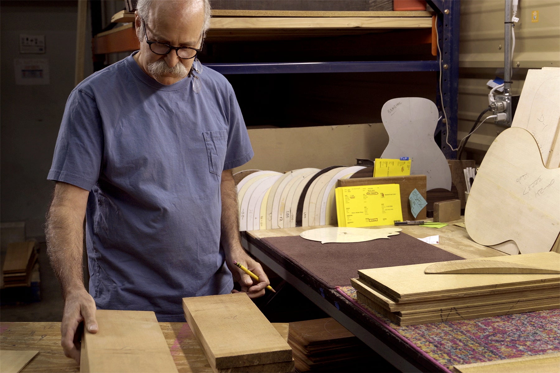 Man in blue shirt inspecting tonewood pieces on a workbench surrounded by guitar body templates and stacked wood sheets.