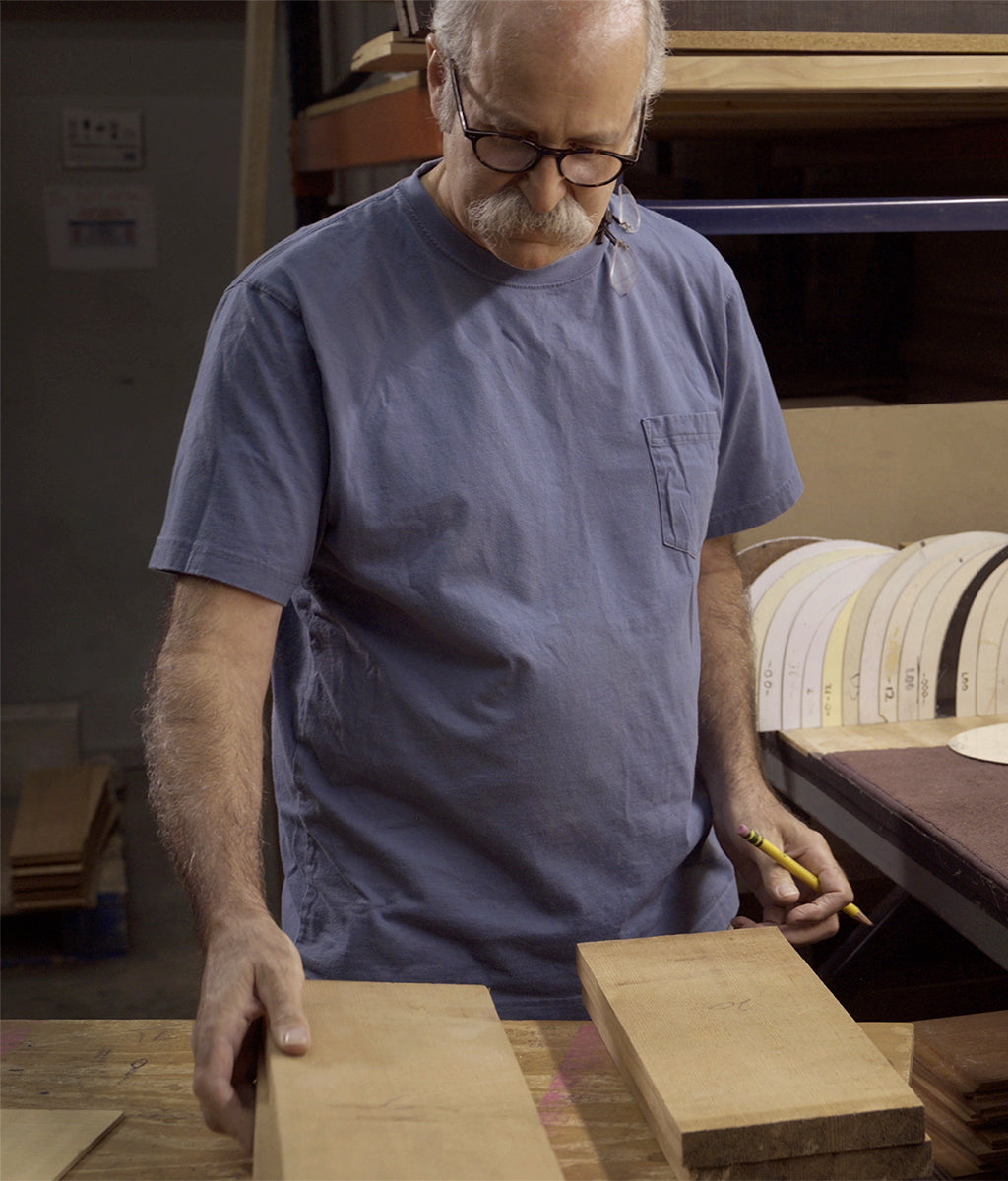 Bruce from Collings Guitars stands at a workbench evaluating two blocks of tonewood, holding one piece in each hand.