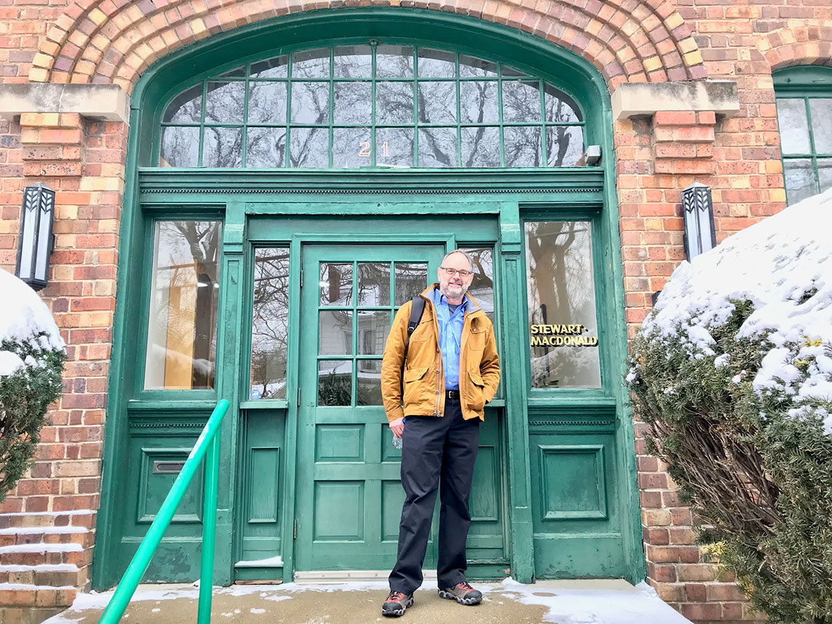 Man in a brown jacket stands smiling in front of a green door on a brick building labeled