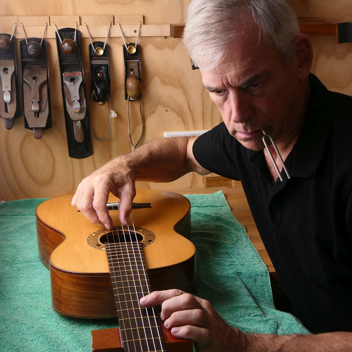 Trevor Gore adjusting a classical guitar on a workbench, with a tuning fork in his mouth and hand tools hanging on the wall behind him.
