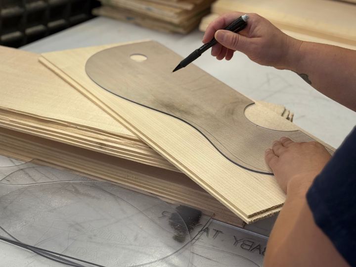 Hands tracing guitar top shapes onto stacked spruce billets for bookmatching and layout alignment.
