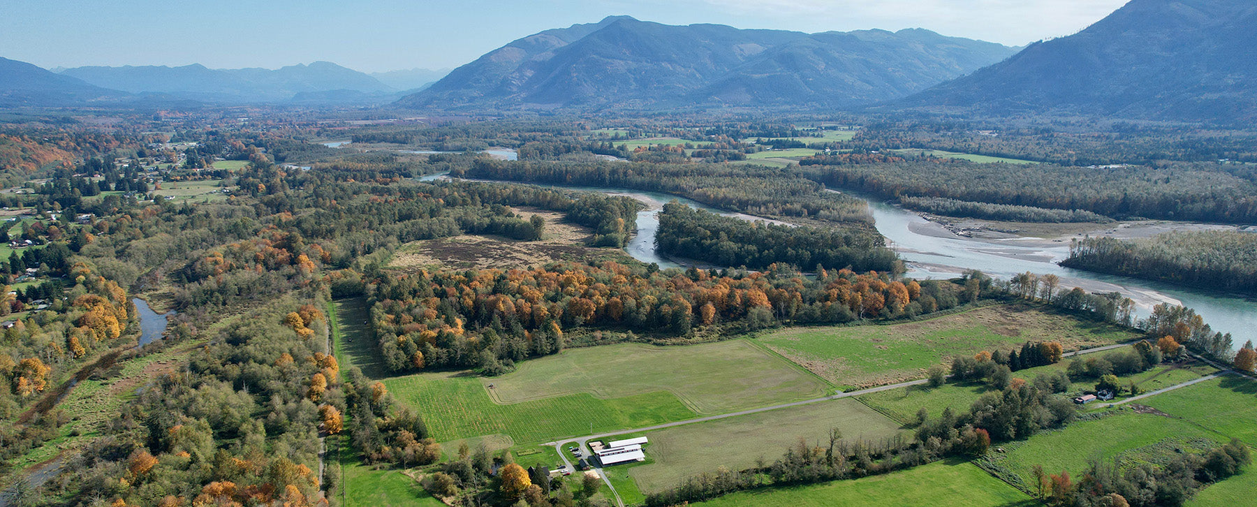 Aerial view of a lush valley with a winding river, surrounded by autumn-coloured forests and distant mountains under a clear blue sky.