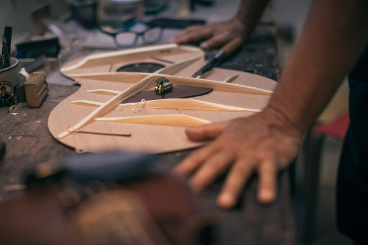 Close-up of a luthier’s hands working on guitar bracing with a chisel and plane tool on a wooden workbench. Spruce top in progress.