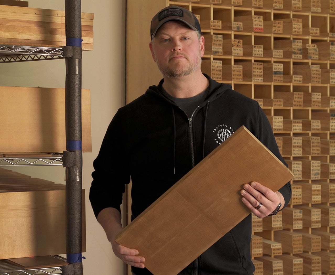 A Pacific Rim Tonewoods employee stands in front of wood inventory shelves, holding a piece of tonewood used in guitar making.