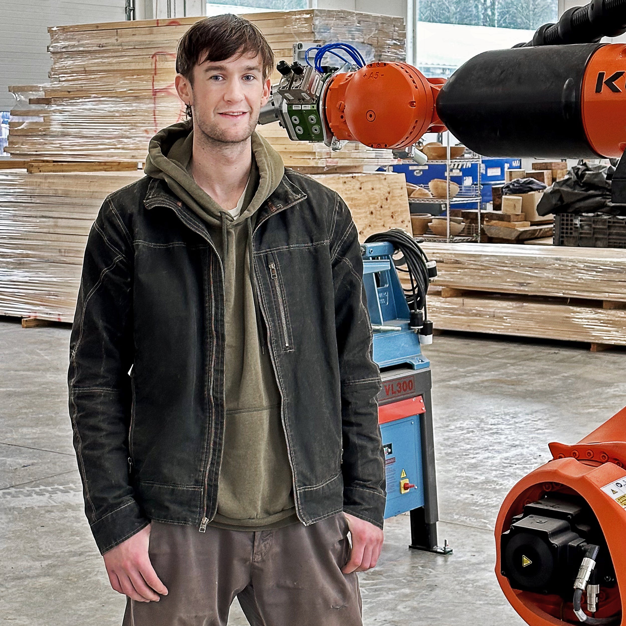 Young man standing in a workshop beside an industrial robotic arm and stacked wood panels.