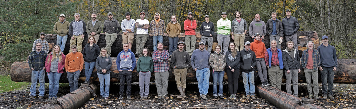 A large group portrait of Pacific Rim Tonewoods staff standing outdoors in front of stacked logs. The team is dressed in work attire, smiling and posed in two rows.