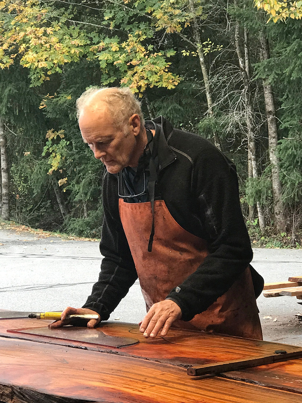 Individual outlines guitar shapes on a slab of figured koa wood. He works outdoors, surrounded by trees in early autumn.