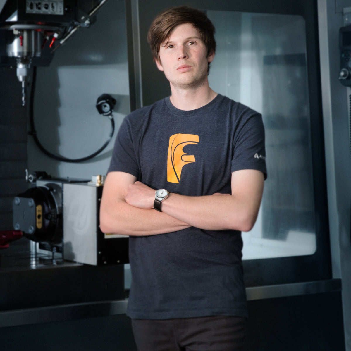 Jake Wells poses confidently in a workshop beside CNC equipment. He wears a dark T-shirt with a stylized “F” and crosses his arms.
