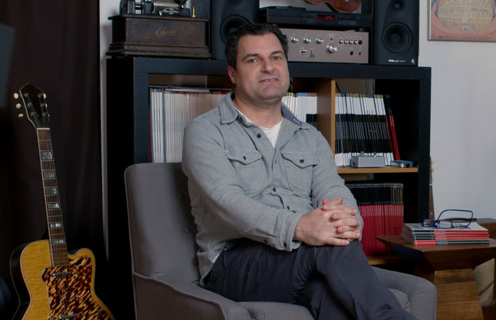 Jason Verlinde is seated in a music-themed room with a semi-hollow electric guitar and a bookshelf of guitar magazines in the background.