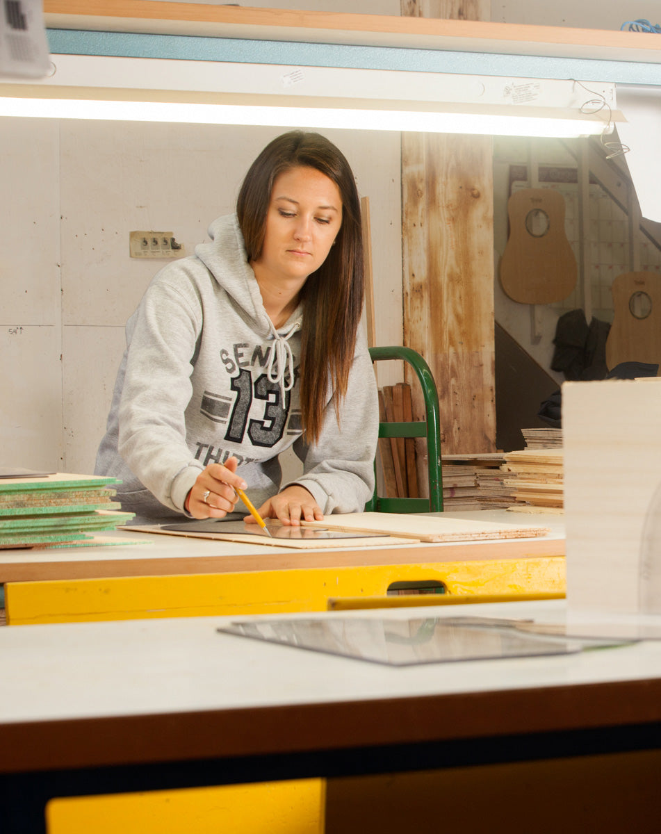 The individual at Pacific Rim Tonewoods workshop carefully traces a design on wood, with guitars hanging in the background.