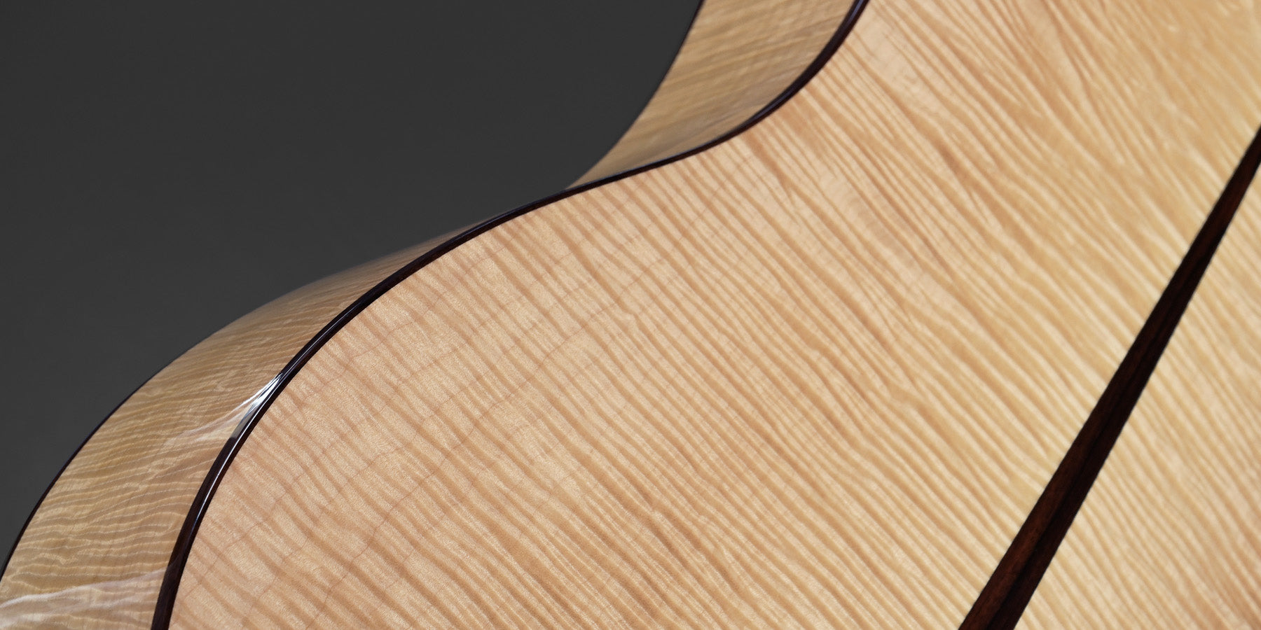 Close-up of a guitar's back, showcasing smooth, light wood with dark edges against a gray background. The wood grain appears sleek and polished.