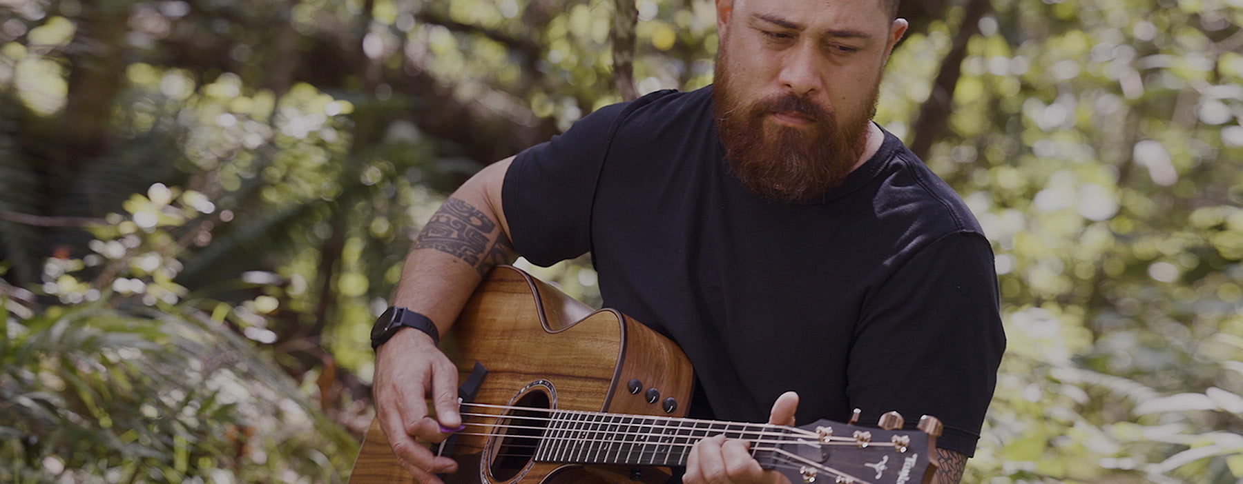 Musician Tavana plays an acoustic guitar in a lush forest setting. Sunlight filters through the trees as he focuses on his performance.